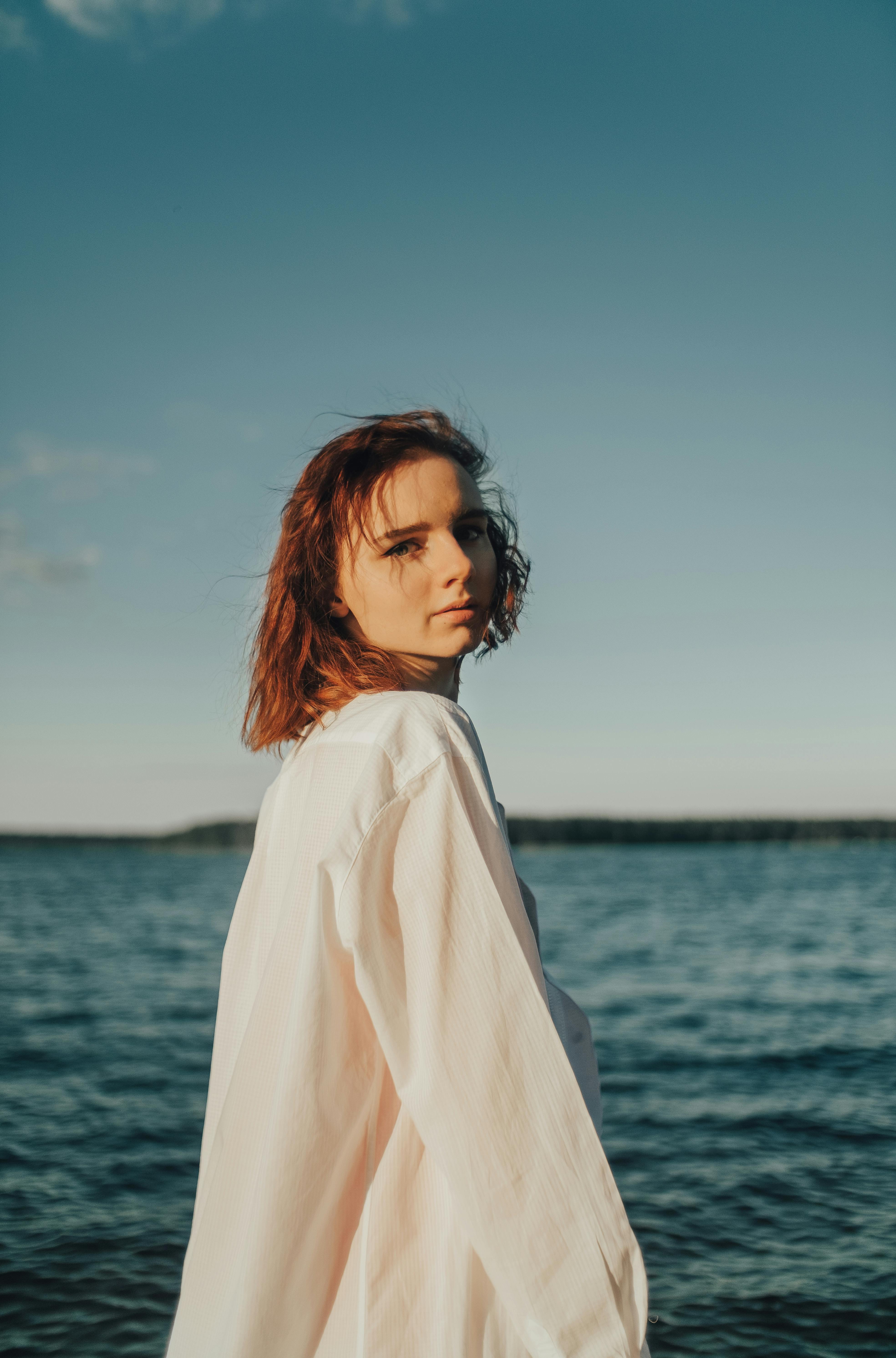 Woman in a white dress standing by a body of water with a clear sky.